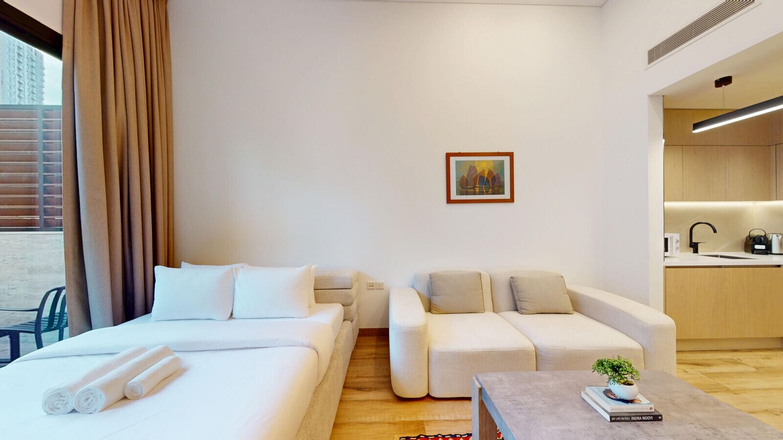Een moderne hotelkamer met twee bedden, een bank, een kleine tafel met een plant en een keukenhoek zichtbaar door een open deur.