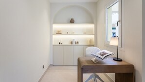 Un couloir avec une alcôve en arc blanc contenant des étagères ornées d’objets décoratifs, une lampe de lecture et un fauteuil avec un livre ouvert.
