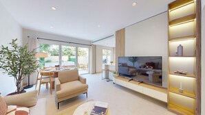 Un salon moderne doté d’un grand téléviseur à écran plat, d’un canapé, d’un coin repas avec une table et des chaises, et d’une grande fenêtre offrant une vue sur l’extérieur.