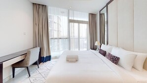 Ein Hotelzimmer mit einem großen Bett, einem Schreibtisch und einem Sessel. Ein Fenster mit Vorhängen bietet Blick auf umliegende Gebäude.