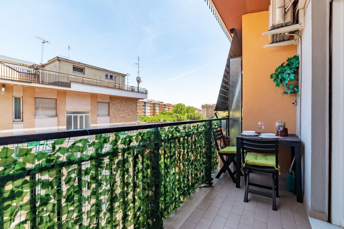 A balcony with a green plant barrier, a table with chairs, and a small window.