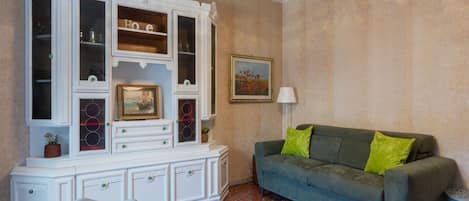 Una habitación con un sofá verde, un armario blanco con puertas de cristal, un cuadro enmarcado y una lámpara.