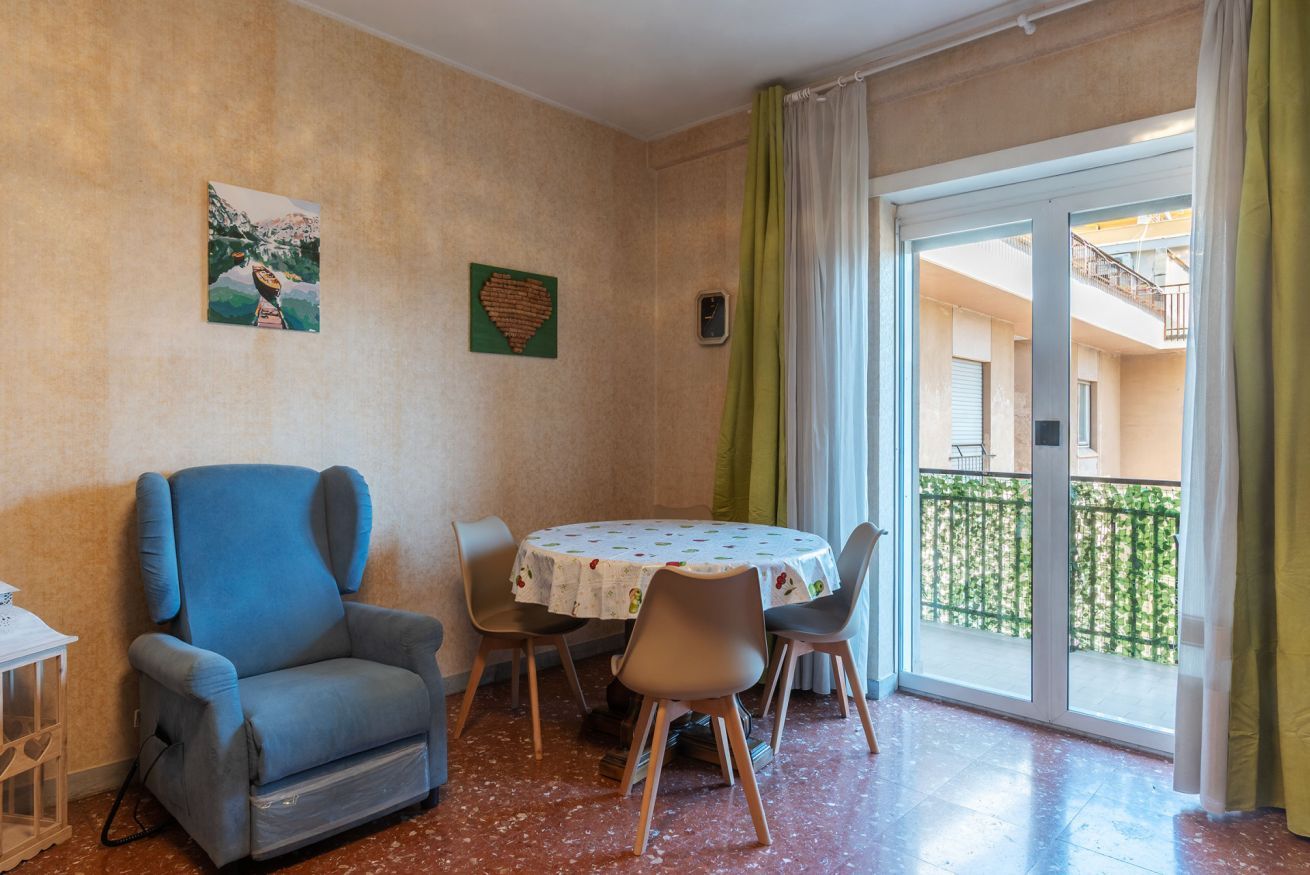 Een kamer met een blauwe fauteuil, een ronde tafel met een tafelkleed met bloemenprint en een uitzicht op een balkon met een groene balustrade.