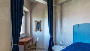 Ein Schlafzimmer mit einem Bett, einem Schreibtisch, einem Stuhl, einem kleinen Tisch mit Lampe und einem Fenster mit Vorhängen.