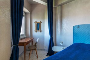 Ein Schlafzimmer mit einem Bett, einem Schreibtisch, einem Stuhl, einem kleinen Tisch mit Lampe und einem Fenster mit Vorhängen.