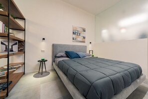 Een moderne slaapkamer met een grijs bed, een boekenkast, een klein rond tafeltje en een kunstwerk aan de muur.