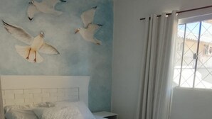 Ein Schlafzimmer mit Bett, weißer Bettwäsche, einem Kopfteil, einem Wandbild von Möwen, einem Fenster mit Vorhängen und einem braunen Teppich.