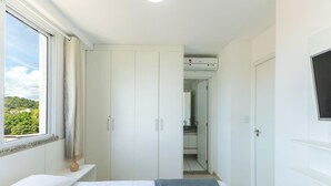 A hotel room with a bed, a wardrobe, a bathroom with a shower, and a view of greenery outside. - Apt w/ Balcony Near the Beach in Ilheus Rpd0301 (Ilhéus)
