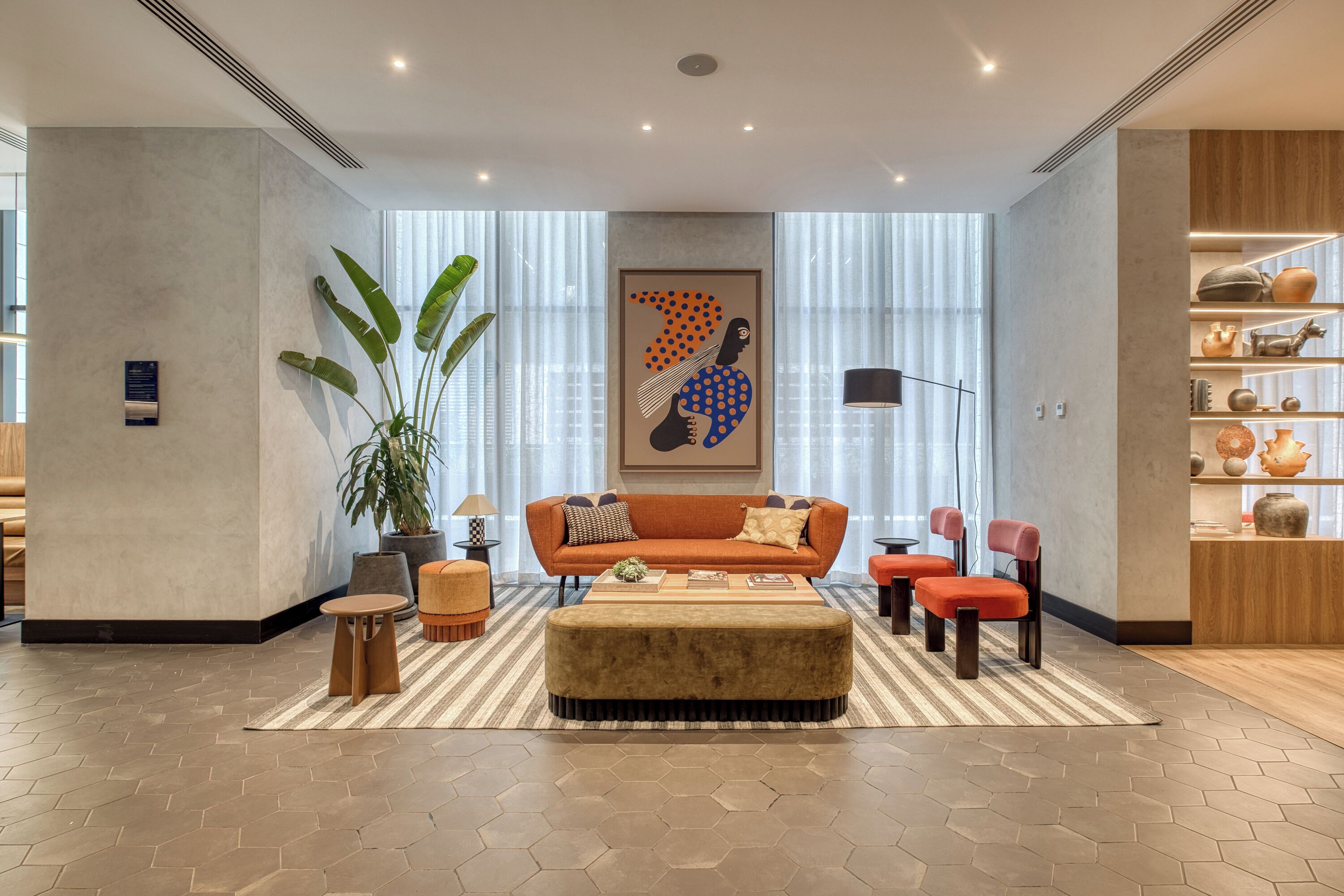A modern living room with a sofa, ottoman, and chairs, a large plant, and a wall with shelves and decorative items.