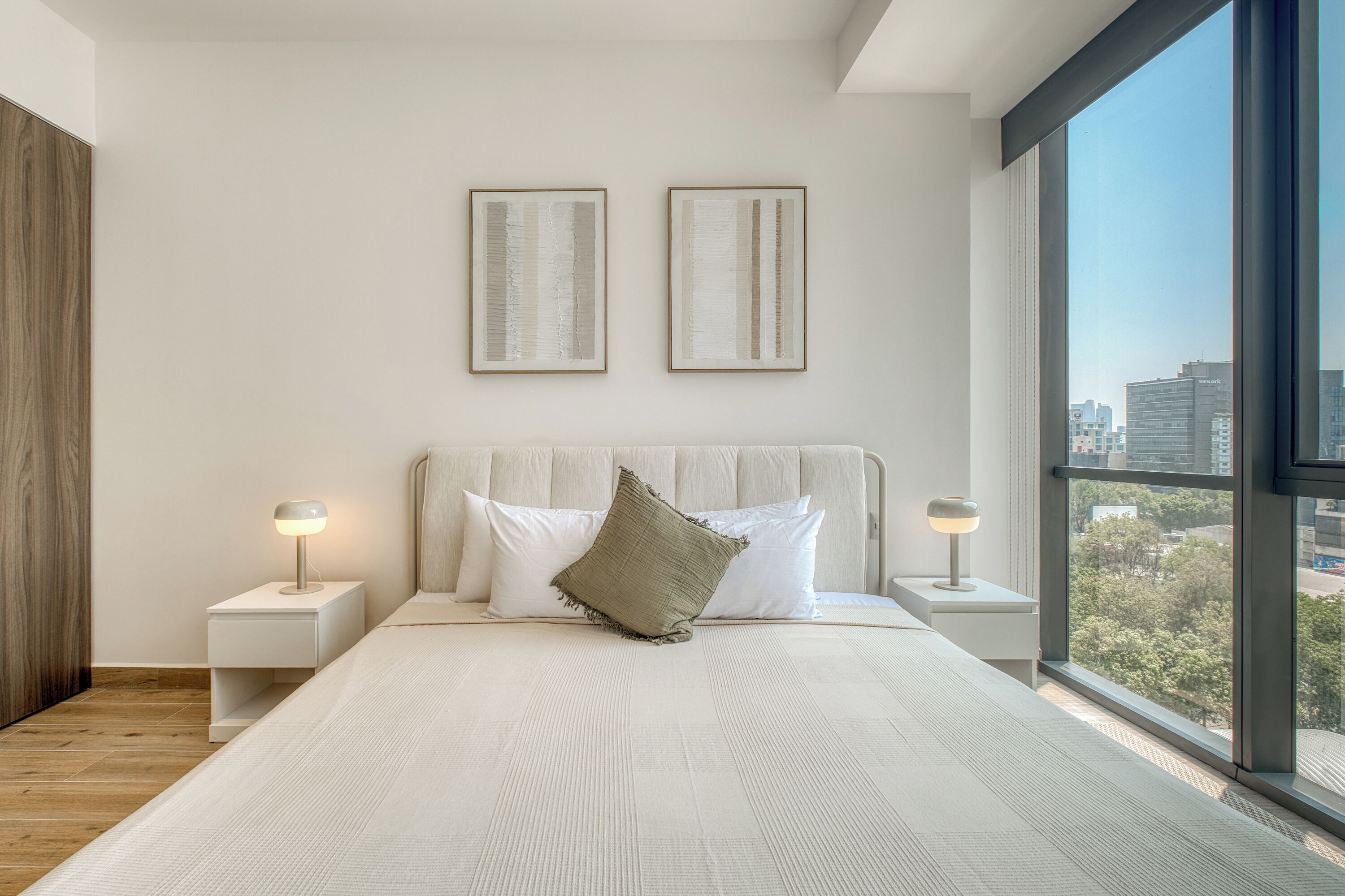 A modern bedroom with a large bed, two bedside tables with lamps, a view of the city through large windows, and two framed artworks on the wall.