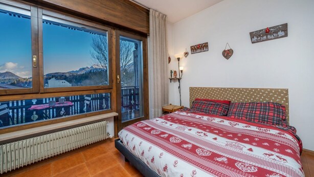 A bedroom with a large window, a bed with patterned bedding, a radiator, and a view of mountains. - GuestHost - In Tarvisio, a cozy 35 sqm studio for 2 guests, located on the second floor of a residential building with an elevator (not suitable for disabled persons). Surrounded by greenery, the building offers an open view of the city center, the sport (Tarvisio)