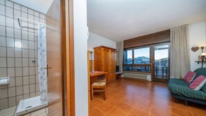 A bathroom with a shower, a wooden cabinet, a chair, and a large window with a view. - GuestHost - In Tarvisio, a cozy 35 sqm studio for 2 guests, located on the second floor of a residential building with an elevator (not suitable for disabled persons). Surrounded by greenery, the building offers an open view of the city center, the sport (Tarvisio)