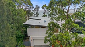 A modern multi-story house with a flat roof, surrounded by dense greenery and overlooking a body of water. - The Villa at Maloneys (Maloneys Beach)