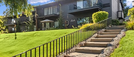 A modern two-story house with a well-maintained lawn, a staircase with a metal railing, and a small garden area with a bench.