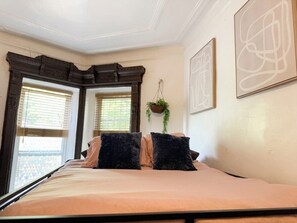 Une chambre à coucher avec un lit, deux fenêtres, une plante suspendue et deux œuvres d’art encadrées accrochées au mur.
