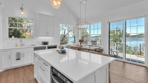 A modern kitchen with a large island, white cabinetry, and large windows overlooking a body of water. - Luxury Lakefront Retreat (Graford)
