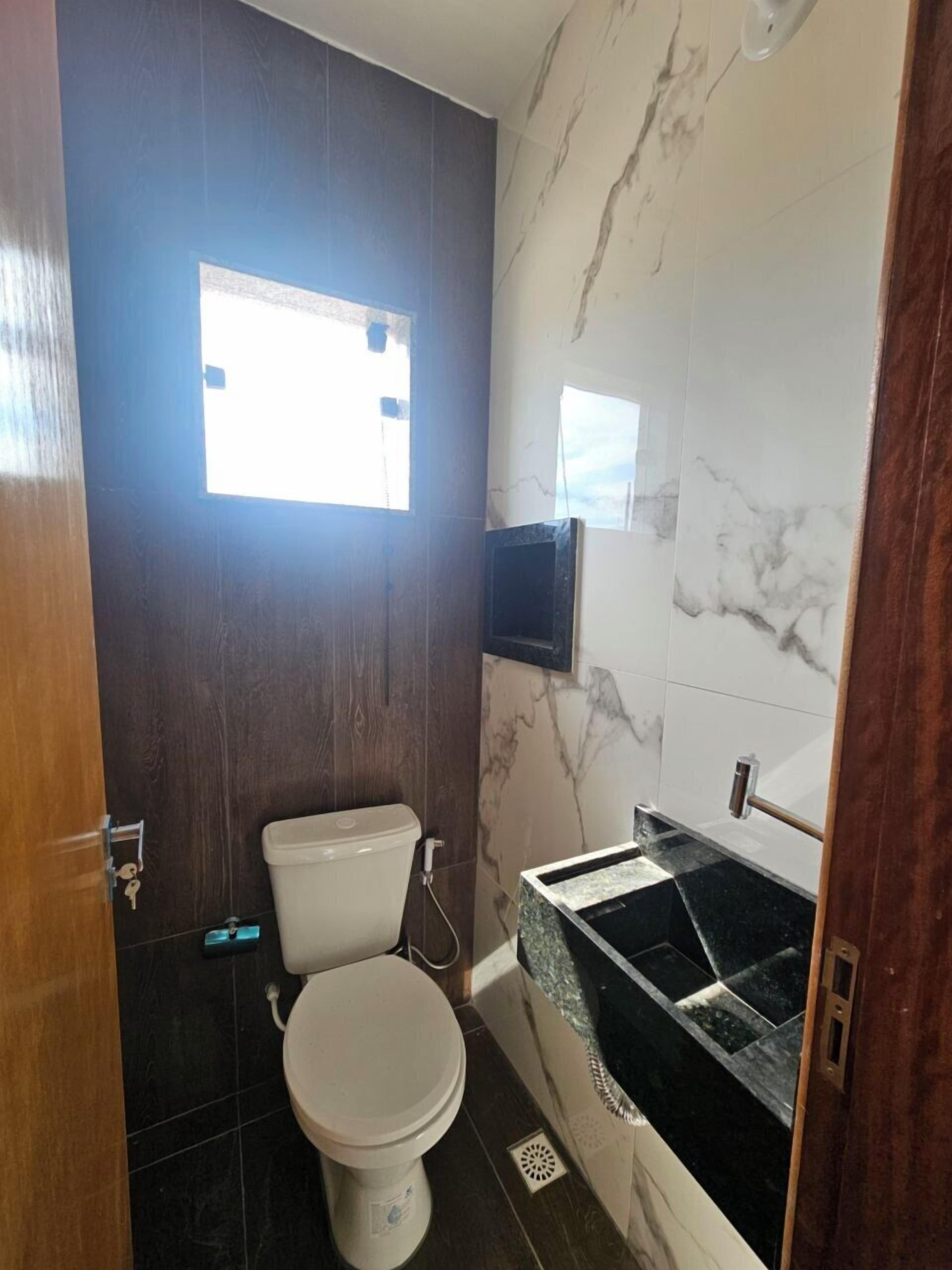 Une salle de bain avec des toilettes, un lavabo et un miroir. Il y a une fenêtre au-dessus des toilettes et un téléviseur fixé au mur.