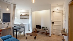 A modern living room with a blue sofa, a glass coffee table, and a dining area with a white table and chairs. There is a kitchen in the background with white cabinets and a white refrigerator. - Anjos City Center Apartment by Homing (Lisbon)