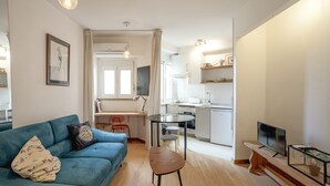A modern living room with a blue sofa, a wooden coffee table, and a TV on a wooden stand. A kitchen is visible in the background with white cabinets and a dining area. - Anjos City Center Apartment by Homing (Lisbon)