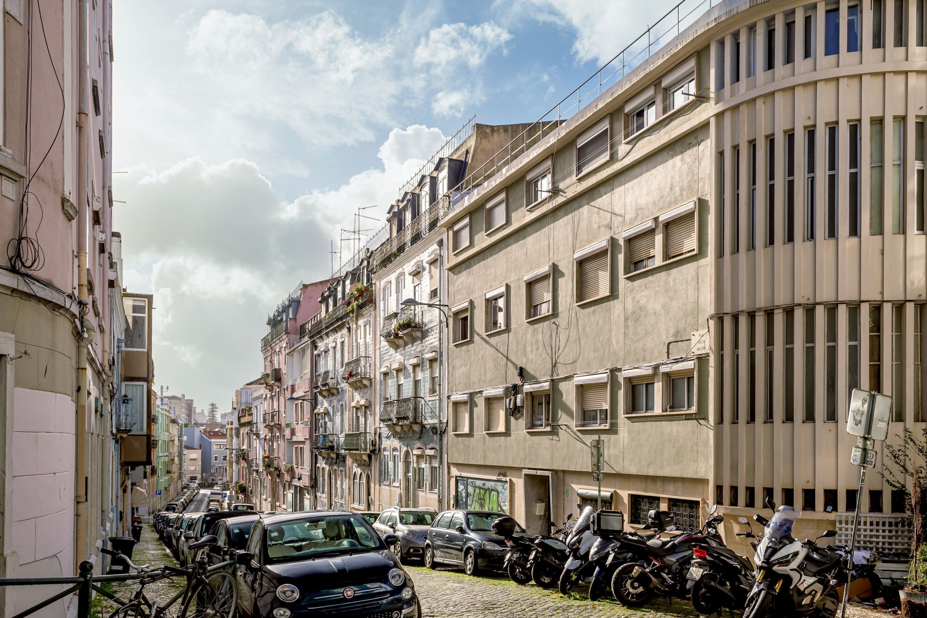Eine schmale Straße, gesäumt von parkenden Autos und Motorrädern, mit Häusern, die Balkone und Fenster haben, und einem klaren Himmel.
