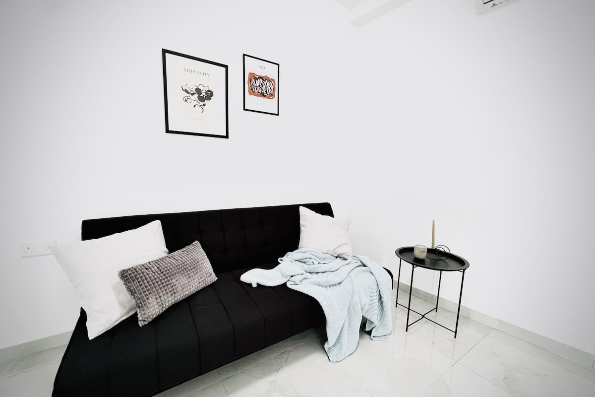 A modern, minimalist bedroom with a black sofa, white pillows, and a small round table with a candle. Two framed artworks are hung on the wall.