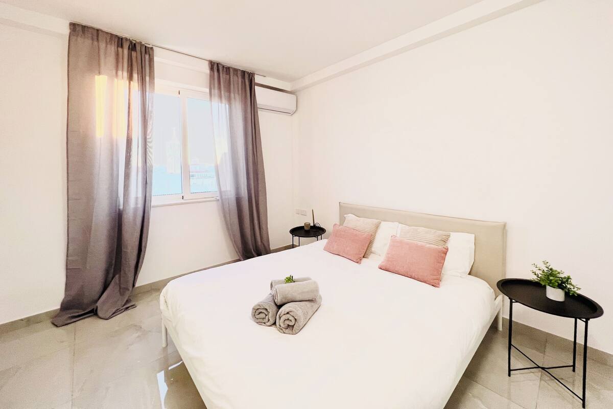 A modern hotel room with a large bed, a small side table, and a window with curtains.