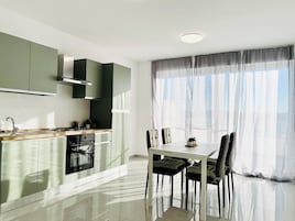 Una cucina moderna con zona pranzo, caratterizzata da un'ampia finestra con tende leggere, un forno a incasso e un tavolo da pranzo con sedie.