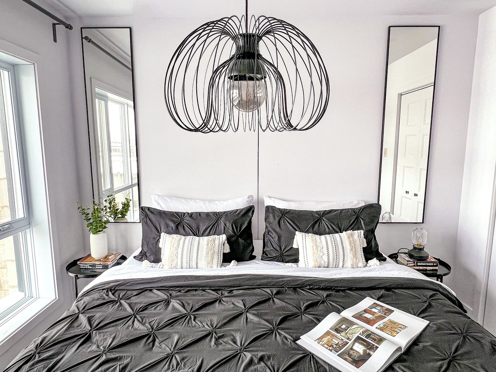 A bedroom with a black upholstered bed, two bedside tables with lamps, a large mirror, and a unique black chandelier.