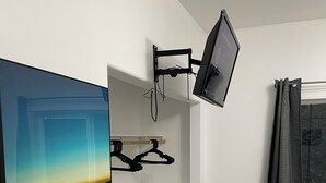 Quarto com televisão na parede, um quadro, um porta-roupas, janela com cortinas e piso de madeira.