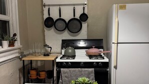 Een kleine keuken met een wit aanrecht, een fornuis, een hangende kruidenrek en een koelkast.