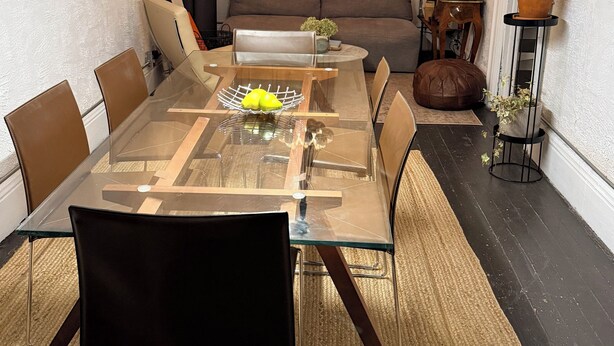 Een eetgedeelte met een tafel met glazen blad, bruine leren stoelen, een geweven vloerkleed en een hanglamp.