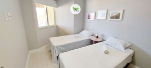 Hotellihuoneessa on kaksi sänkyä, pieni pyöreä pöytä, jossa on lamppu, sekä kolme kehystettyä kuvaa seinällä.