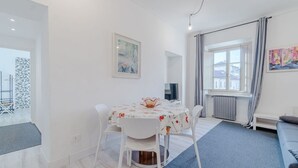 Ein modernes Wohnzimmer mit Esstisch, weißen Stühlen, einem blauen Teppich, einem grauen Sofa und einem Fenster mit Vorhängen.