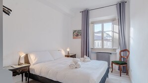 Ein Schlafzimmer mit einem Bett, Nachttischen, einem Sessel, einem Fenster mit Vorhängen und einer Heizung.