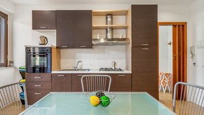 A modern kitchen with dark cabinets, a stainless steel range hood, and a glass dining table with a fruit bowl. - GuestHost - Cozy 43 sqm ground floor apartment, perfect for three people (accessible via a few steps).The property enjoys a prime location, directly on the beach, with convenient access to the sea. The Taormina - Giardini train station is less than a 10- (Naxos)