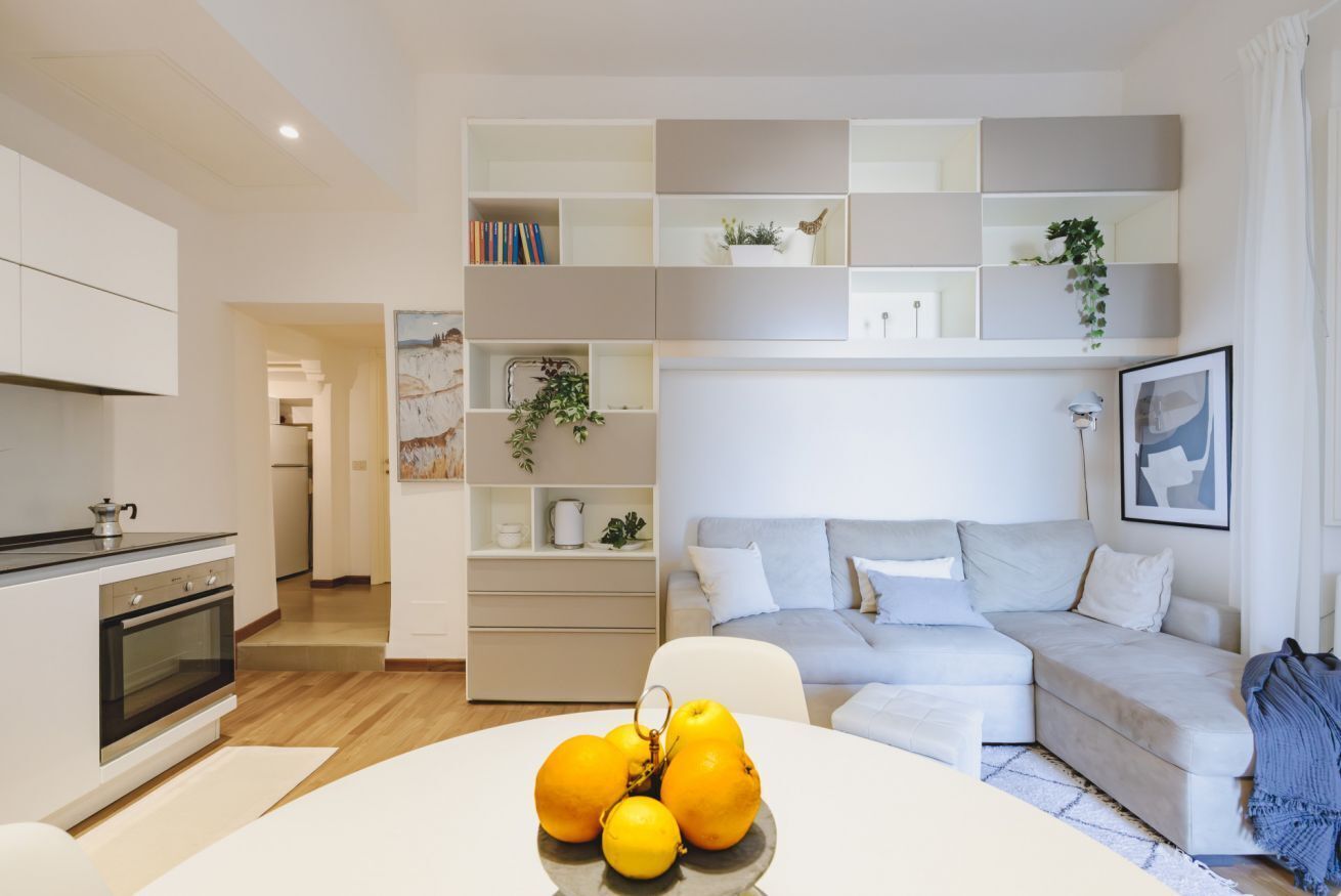 A modern living room with a sofa, a dining table with fruit, and built-in shelving with books and decorative items.