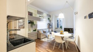 Una cocina moderna con un lavamanos negro, una zona de comedor con una mesa redonda y sillas blancas, un sofá gris y una lámpara colgante blanca.