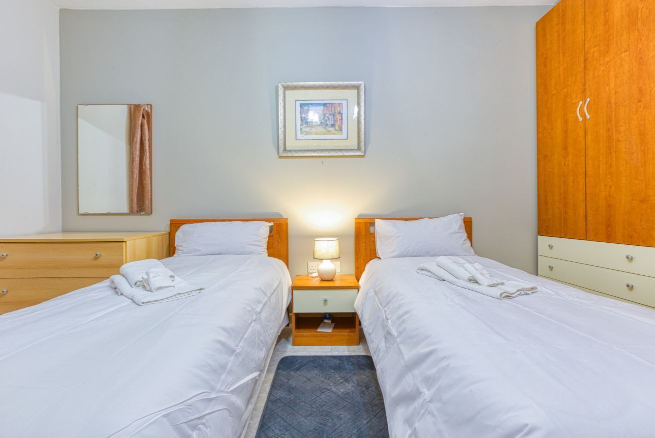 Habitación de hotel con dos camas, una mesita de noche con lámpara, un armario de madera, un espejo y un cuadro en la pared.