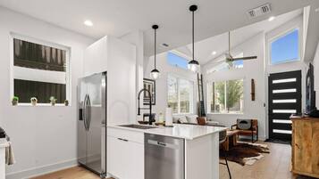 Una cocina moderna con una isla central, electrodomésticos de acero inoxidable, iluminación colgante y un comedor con mesa y sillas de madera.
