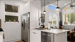 Una cocina moderna con un refrigerador de acero inoxidable, lavavajillas y una isla grande con pileta. Hay plantas en macetas en el alféizar de la ventana y vistas al exterior a través de las ventanas.