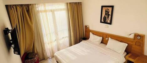 A hotel room with a large bed, a wooden headboard, a bedside table, a TV mounted on the wall, and a small red chair.