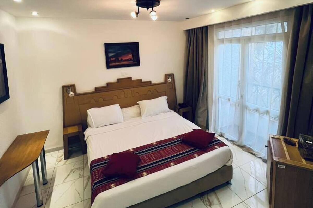 A hotel room with a large bed, a wooden headboard, a bedside table, a desk, and a window with curtains.