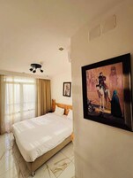 A hotel room with a bed, a ceiling light, a window with curtains, and a framed picture on the wall.