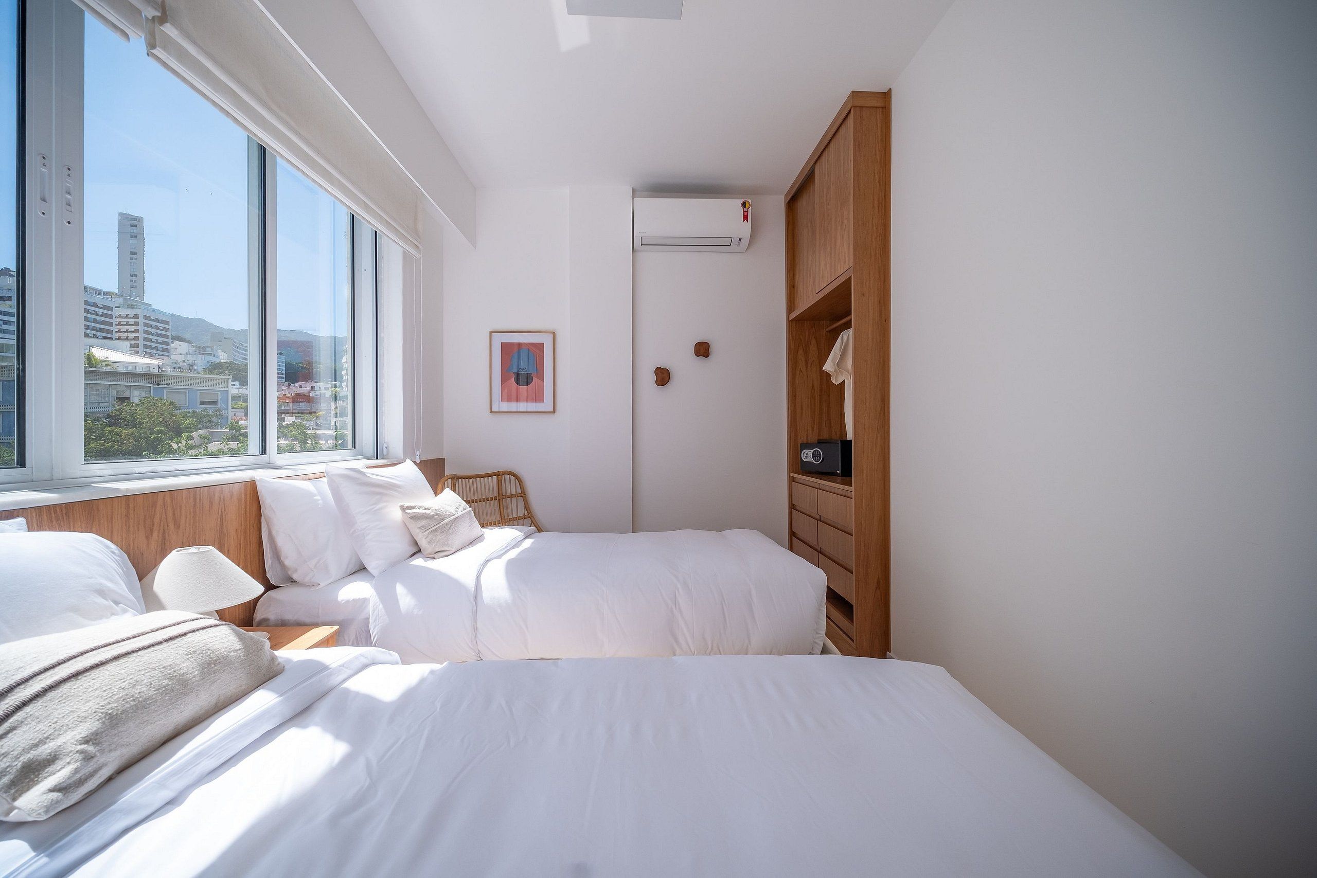 A hotel room with two beds, a wooden wardrobe, an air conditioning unit, and a window with a city view.