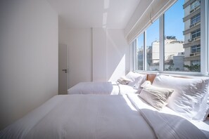 Een lichte hotelkamer met een groot raam, een bed met witte lakens en uitzicht op de stad.