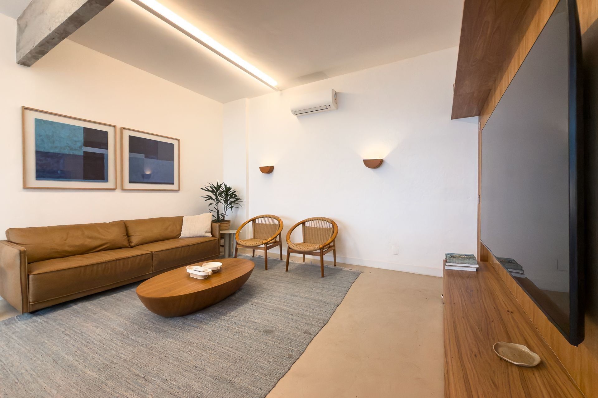 A modern living room with a brown sofa, two wicker chairs, a coffee table, and a flat-screen TV mounted on the wall.