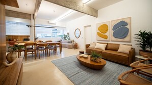 Una sala de estar moderna con sofá, mesa de centro y sillas, un comedor con mesa y sillas, y una cocina al fondo.