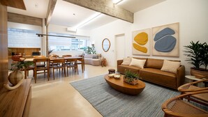 Un salon moderne avec un canapé, une table basse et des fauteuils, un coin repas avec une table et des chaises, et une cuisine en arrière-plan.
