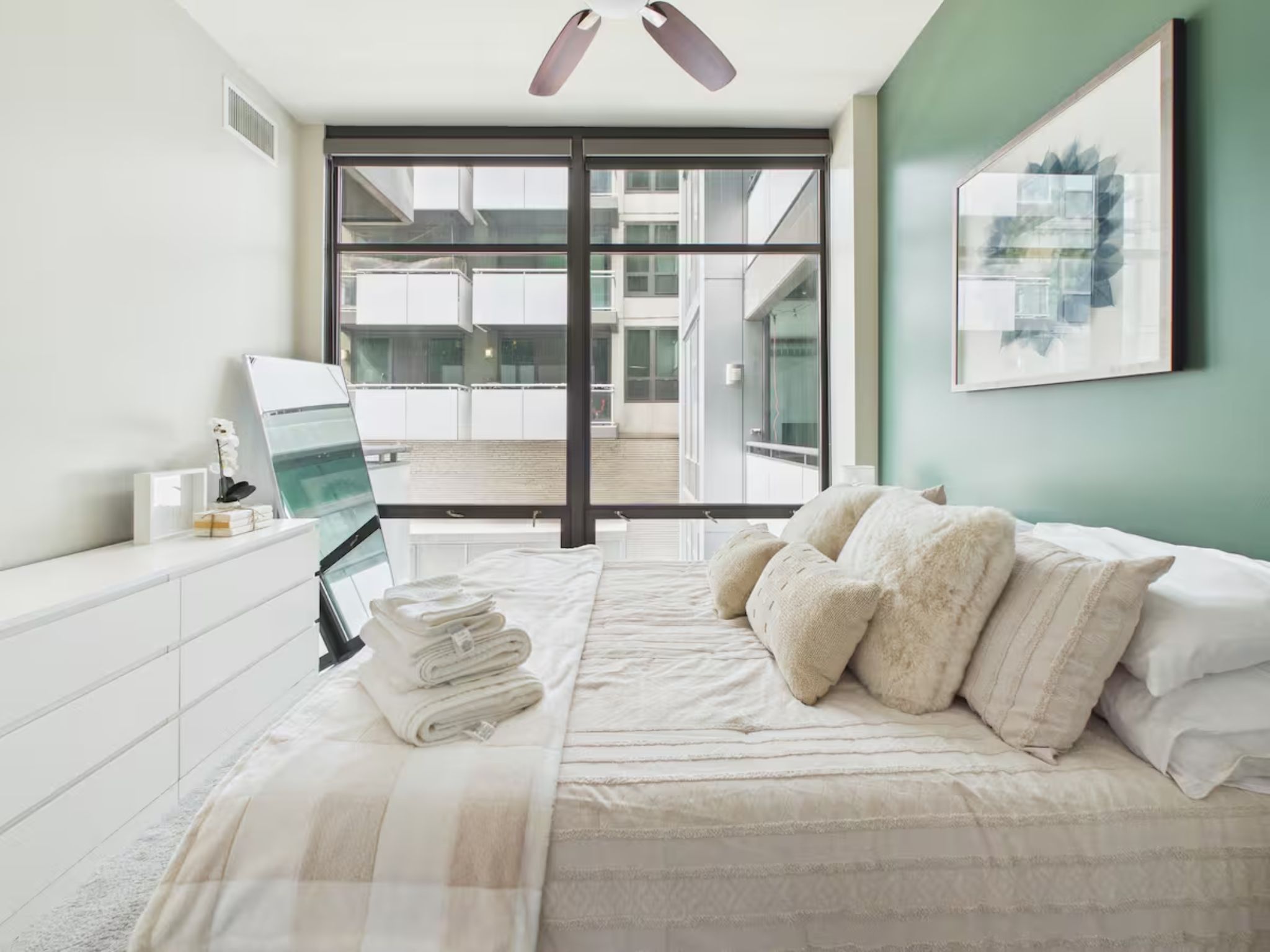 A bedroom with a bed, a bedside table, a window with a view of buildings, and a ceiling fan.