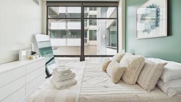 A bedroom with a bed, a bedside table, a window with a view of buildings, and a ceiling fan.